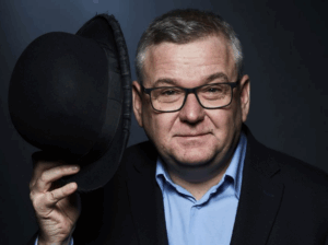 Na zdjęciu znajduje się uśmiechnięty mężczyzna w okular. Jest on ubrany w błękitną koszulę oraz granatową marynarkę. W swej prawej dłoni trzyma czarny kapelusz.