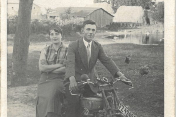 Czarno-białe zdjęcie przedstawia mężczyznę i kobietę siedzących na motocyklu. Mężczyzna, ubrany elegancko w garnitur i krawat, trzyma kierownicę motocykla, a kobieta siedzi za nim bokiem, z rękami skrzyżowanymi na piersi. Ma na sobie spódnicę i koszulę bez rękawów w kratkę. W tle widać wiejski krajobraz: drzewa, staw, zabudowania gospodarcze oraz stogi siana. Na ziemi w pobliżu chodzą kury. Numer rejestracyjny motocykla to „PZ 4253”. Zdjęcie ma lekko zaokrąglone rogi i nosi ślady starzenia.