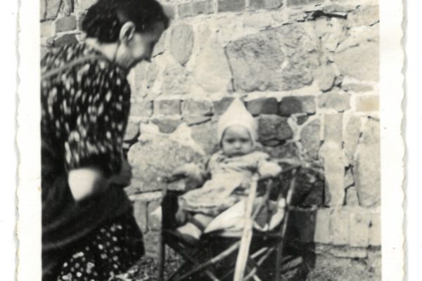 Czarno-białe zdjęcie przedstawiające starszą kobietę w ciemnej sukience w kwiaty, pochyloną w kierunku małego dziecka siedzącego na krzesełku. Kobieta znajduje się po lewej stronie kadru, odwrócona częściowo tyłem. Dziecko ma na sobie jasne ubranko i czapeczkę, siedzi w prostym, drewnianym krzesełku z podpórkami, opartym o kamienny mur z widocznymi cegłami. Zdjęcie zostało zrobione na zewnątrz, prawdopodobnie na podwórku lub w ogrodzie. Całość utrzymana w ciepłej, domowej atmosferze lat 40. XX wieku.