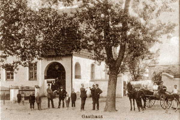 Stare, sepia-tonowane zdjęcie przedstawia scenę sprzed zajazdu (gospody) oznaczonego napisem „Gasthaus” oraz imieniem i nazwiskiem właściciela „Gustav Radeck”. Budynek ma charakterystyczny łukowy portal wejściowy i dwuspadowy dach, a przed nim znajduje się grupa ludzi. Na pierwszym planie widać kilkunastu mężczyzn i chłopców stojących i rozmawiających ze sobą. Część z nich nosi kapelusze i garnitury. W tle, po prawej stronie, stoi zaprzęg konny z powozem — siedzi w nim kobieta w kapeluszu oraz mężczyzna powożący. Obok koni stoją dzieci i dorośli. Scena ma charakter codzienny, być może jest to moment towarzyskiego spotkania lub początek jakiegoś wydarzenia. Zdjęcie prawdopodobnie pochodzi z przełomu XIX i XX wieku i ukazuje fragment lokalnego życia społecznego.