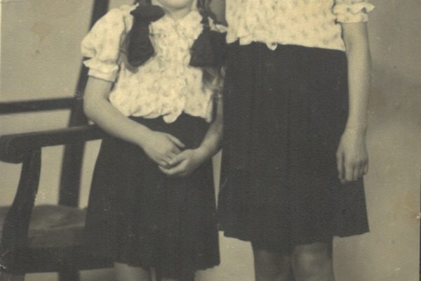 Na czarno-białym, lekko zniszczonym zdjęciu portretowym widoczne są dwie dziewczynki stojące obok siebie. Obie mają na sobie identyczne stroje – jasne bluzki z bufkami i wzorkiem oraz ciemne spódniczki. Włosy mają związane w warkocze z dużymi, ciemnymi kokardami przy głowach. Dziewczynka po lewej jest młodsza i niższa, stoi lekko przygarbiona, z rękoma splecionymi z przodu. Druga, starsza i wyższa, stoi prosto, z rękami luźno opuszczonymi wzdłuż ciała. Obie mają białe skarpetki i klasyczne buciki. W tle widoczny jest prosty, drewniany fotel z obitym siedziskiem. Zdjęcie wykonane jest w atelier fotograficznym na neutralnym tle.