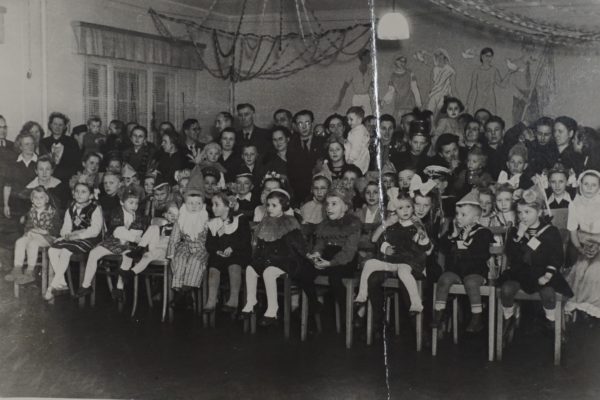 Czarno-białe zdjęcie przedstawia dużą grupę dzieci i dorosłych zgromadzonych w sali, prawdopodobnie podczas jakiejś uroczystości lub przedstawienia. W pierwszym rzędzie siedzą dzieci w różnorodnych strojach – niektóre ubrane są w kostiumy postaci z bajek, inne w stroje ludowe lub eleganckie ubrania. Część dzieci ma na głowach czapki, korony lub kokardy. Za dziećmi stoją dorośli – kobiety i mężczyźni – uśmiechający się i obserwujący wydarzenie. W tle widać ściany sali ozdobione łańcuchami i malowidłami przedstawiającymi ludzi i gołębie. Cała scena ma radosny, uroczysty charakter, typowy dla szkolnych lub przedszkolnych wydarzeń sprzed wielu lat.