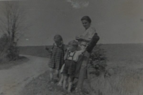 Czarno-białe zdjęcie przedstawia kobietę z trójką dzieci na wiejskiej drodze. Kobieta stoi z najmłodszym dzieckiem zawiniętym w becik, które trzyma w ramionach. Obok niej stoi chłopiec w krótkich spodenkach i szelkach, a z lewej strony dziewczynka w sukience w kratkę. Wszyscy stoją przy skraju drogi gruntowej, która zakręca w tle i prowadzi przez wiejski krajobraz. W tle widać otwarte pola i pojedyncze drzewa. Zdjęcie oddaje atmosferę spokojnego, rodzinnego spaceru na wsi w słoneczny dzień.