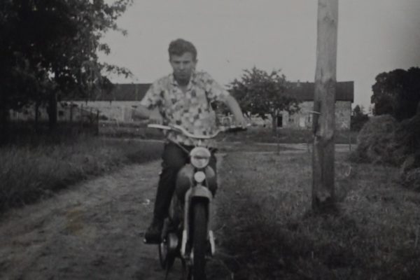 Czarno-białe zdjęcie przedstawia młodego mężczyznę jadącego motorowerem po wiejskiej, nieutwardzonej drodze. Mężczyzna ma na sobie koszulę z krótkim rękawem w geometryczne wzory i ciemne spodnie. Tło stanowi wiejski krajobraz: pola, drzewa, drewniany słup oraz zabudowania z kamienia, prawdopodobnie stodoły lub domy. Po prawej stronie kadru widać kopiec siana. Zdjęcie uchwycone jest w ruchu, z perspektywy na wprost, co daje dynamiczne wrażenie.