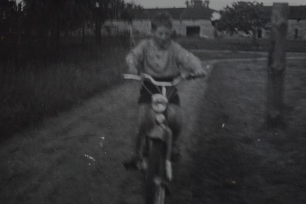 Czarno-białe, lekko rozmazane zdjęcie przedstawia chłopca jadącego na motorowerze po wiejskiej drodze. Chłopiec ubrany jest w jasną bluzę i krótkie spodenki. W tle widoczne są pola, drzewa oraz zabudowania gospodarcze – stodoły lub domy z kamienia.