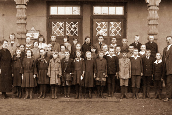 Stare zdjęcie w sepii przedstawia dużą grupę dzieci oraz dwóch dorosłych (kobietę i mężczyznę) nauczycieli, stojących przed wejściem do budynku szkolnego. Grupa liczy ponad 30 osób i została ustawiona w trzech rzędach – dzieci z przodu stoją na ziemi, w środkowym rzędzie stoją nieco wyżsi uczniowie, a z tyłu ustawiono najstarsze dzieci i dorosłych. Dzieci ubrane są w proste, skromne ubrania z początku XX wieku – dziewczynki w sukienki i buty sznurowane, chłopcy w garniturki, marynarki lub płaszcze. Niektóre dzieci trzymają ręce z tyłu lub z przodu ciała, pozując poważnie do zdjęcia. Na tle widać dwa duże, drewniane drzwi z oszklonymi okienkami w kształcie rombów. Po bokach drzwi znajdują się ozdobne kolumny. Atmosfera zdjęcia jest formalna – to typowe zdjęcie klasowe zrobione prawdopodobnie w okresie międzywojennym przed budynkiem szkoły w Chludowie.