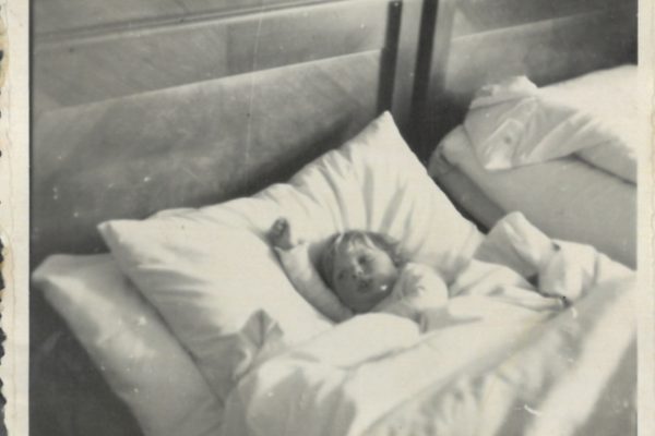 Czarno-białe zdjęcie przedstawia małe dziecko leżące w łóżku, otulone grubą pościelą. Dziecko ma jasne włosy i wygląda, jakby miało może rok lub dwa. Główka dziecka opiera się na dużej poduszce, a rączki wystają spod kołdry – jedna z nich uniesiona w górę. Na twarzy dziecka widać spokojny, nieco zaspany wyraz, jakby właśnie się obudziło lub miało zasypiać. W tle widać drugie łóżko, również starannie pościelone, oraz drewniane zagłówki obu łóżek. Całość sprawia wrażenie wnętrza prostego, schludnego pokoju – być może w domu rodzinnym lub sanatorium. Zdjęcie ma ramkę z fotorożkami, co sugeruje, że było przechowywane w albumie.