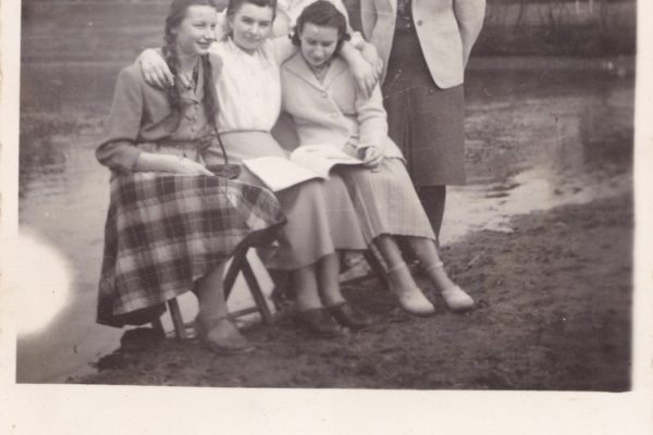 Czarno-białe zdjęcie przedstawia pięć młodych kobiet pozujących nad brzegiem chludowskiego Balatonu. Trzy z nich siedzą na małych krzesełkach, dwie stoją za nimi, wszystkie ubrane są w skromne, eleganckie stroje z lat 40. lub 50. XX wieku. Kobiety siedzące trzymają otwartą książkę, nad którą się pochylają, co może sugerować wspólną lekturę lub naukę. Jedna z nich ma warkocze i nosi kraciastą spódnicę. Kobiety stojące obejmują siedzące ramionami – na ich twarzach widać życzliwość i bliskość. W tle widać bezlistne drzewa oraz wiejskie domy z dwuspadowymi dachami, co sugeruje, że zdjęcie wykonano wczesną wiosną lub późną jesienią. Kadr uchwyca ciepły moment przyjaźni i wspólnoty.
