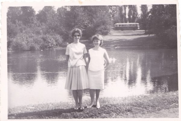 Czarno-białe zdjęcie przedstawia dwie młode kobiety stojące nad brzegiem chludowskiego Balatonu. Obie ubrane są w eleganckie, letnie stroje z lat 60. – kobieta po lewej ma na sobie rozkloszowaną, plisowaną spódnicę i bluzkę z krótkim rękawem, a ta po prawej – prostą sukienkę bez rękawów. Obie uśmiechają się, a kobieta po lewej nosi ciemne okulary przeciwsłoneczne. W tle, po drugiej stronie wody, znajduje się porośnięta drzewami skarpa. Na jej szczycie jedzie autobus – widoczny tylko częściowo, ale wyraźny dzięki kontrastowi z jasnym niebem i koronami drzew. Powierzchnia wody spokojna, z widocznym odbiciem drzew. Zdjęcie ma białą ramkę i lekko postrzępione krawędzie, co świadczy o jego wieku. Panuje pogodna, letnia atmosfera.