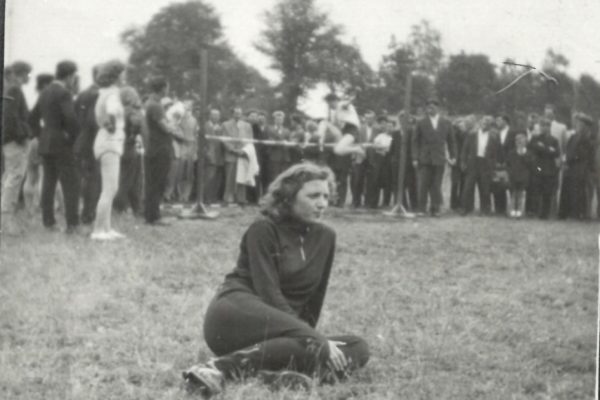 Czarno-białe zdjęcie ukazuje kobietę w sportowym stroju siedzącą na trawie na pierwszym planie, z nogami zgiętymi pod sobą. Jest ubrana w dres, a na nogach ma sportowe buty. Kobieta patrzy w bok, a jej wyraz twarzy wydaje się być skupiony lub zamyślony. W tle znajduje się tłum ludzi stojących wokół prowizorycznego miejsca zawodów lekkoatletycznych — najprawdopodobniej konkursu skoku wzwyż. Można zauważyć poprzeczkę oraz zawodniczkę w białym stroju gimnastycznym w trakcie próby skoku. Zebrani widzowie to głównie mężczyźni w garniturach i młodzież, którzy uważnie obserwują wydarzenie. W oddali widać drzewa, co sugeruje, że zawody odbywają się na otwartym terenie, prawdopodobnie w parku lub na boisku. Atmosfera zdjęcia wskazuje na lokalną imprezę sportową z połowy XX wieku, z dużym zainteresowaniem społeczności lokalnej.