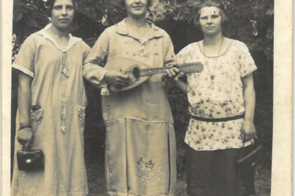 Czarno-białe zdjęcie przedstawia trzy kobiety stojące obok siebie na tle drzew. Kobieta po lewej stronie ma krótkie włosy, nosi sukienkę z kołnierzykiem i trzyma małą torebkę. Kobieta w środku uśmiecha się, ubrana jest w ozdobioną sukienkę z haftem i trzyma mandolinę (lub podobny instrument strunowy). Kobieta po prawej stronie ma krótko ścięte włosy, luźną bluzkę w kwiaty i ciemną spódnicę, również trzyma torebkę. Wszystkie panie mają na sobie ciemne pończochy i eleganckie buty. Zdjęcie ma charakter pamiątkowy, z okresu międzywojennego.