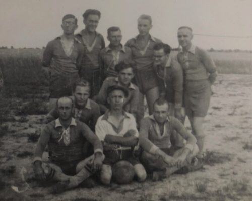 Grupowe zdjęcie drużyny piłkarskiej złożonej z jedenastu mężczyzn. Siedzą i stoją na otwartym polu, kilku z nich w koszulkach z kołnierzykami, jeden trzyma piłkę. Ubrania są różnorodne, niektóre z apaszkami.