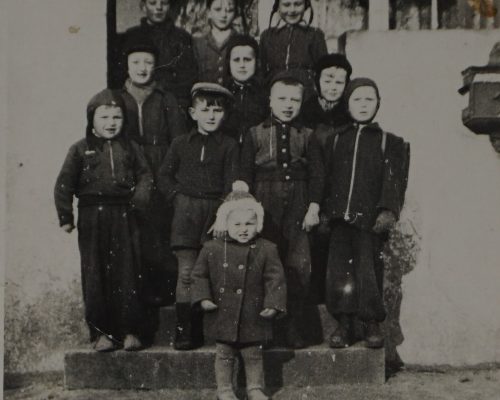 Czarno-białe zdjęcie przedstawia grupę dzieci ustawioną w kilku rzędach na schodkach przed budynkiem. W sumie widać jedenaścioro dzieci — najmniejsze dziecko stoi na najniższym stopniu z przodu, ubrane w ciepły płaszczyk z pomponem na czapce. Pozostałe dzieci są ustawione w dwóch rzędach na schodach i ostatnim rzędzie na górze, tuż przy ścianie budynku. Dzieci ubrane są w zimowe stroje – płaszcze, czapki, szaliki – co sugeruje chłodną porę roku. W tle widać ścianę domu z dużym oknem, przez które można dostrzec firanki i elementy wnętrza.