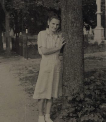 Czarno-białe zdjęcie przedstawia młodą kobietę stojącą na ścieżce obok dużego drzewa. Kobieta ubrana jest w jasną sukienkę z krótkim rękawem i białe buty. Delikatnie opiera się o pień drzewa, lekko przechylona, i uśmiecha się w stronę fotografa. W tle widoczny jest ogrodzony teren, prawdopodobnie cmentarz, z kilkoma pomnikami — w tym jeden większy, przypominający figurę postaci. Wokół drzewa rośnie niska roślinność, a w oddali ciągnie się aleja drzew.