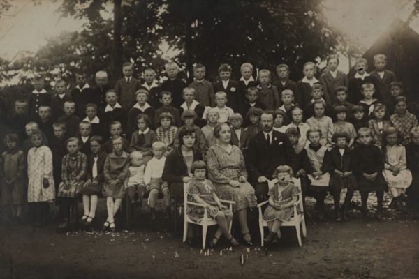 Czarno-białe zdjęcie grupowe przedstawia dużą liczbę dzieci oraz kilku dorosłych. Zdjęcie zostało wykonane na zewnątrz, prawdopodobnie w otoczeniu drzew lub parku, przy naturalnym świetle. Dzieci są ubrane w eleganckie stroje z epoki, głównie sukienki, koszule i marynarki, niektóre z dużymi kołnierzykami. W środkowej części zdjęcia, w pierwszym rzędzie, siedzi troje dorosłych – dwie kobiety i jeden mężczyzna – oraz dwie dziewczynki w małych krzesełkach dziecięcych. Za nimi ustawiono dzieci w kilku rzędach, patrzące prosto w obiektyw. Zdjęcie może pochodzić z lat 20. lub 30. XX wieku