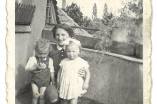 Czarno-białe zdjęcie przedstawia trzy osoby – młodą kobietę i dwoje małych dzieci – pozujące razem na tarasie lub balkonie przy budynku. Kobieta kuca między dziećmi, obejmując dziewczynkę z prawej strony. Chłopczyk po lewej trzyma coś w ręce i patrzy w dół. Dziewczynka ma jasne włosy i dużą kokardę na głowie, ubraną w jasną sukienkę i buciki. Chłopczyk nosi krótkie spodenki na szelkach i długie skarpetki. Za nimi widać dachy z dachówki i fragment okna z daszkiem, a w tle – drzewa i niebo. Zdjęcie ma postrzępione brzegi i delikatne zabrudzenia, co sugeruje jego wiek – z lat 50. XX wieku.