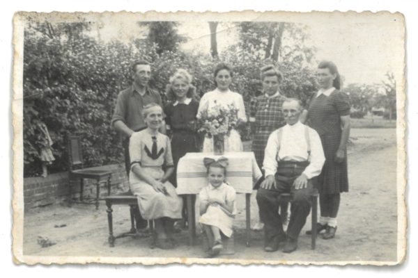 Zdjęcie czarno-białe z 1947 roku przedstawia ośmioosobową grupę ludzi oraz jedno dziecko. Zdjęcie zrobione na zewnątrz, najprawdopodobniej w wiejskim ogrodzie lub na podwórzu przy domu (w tle widać żywopłot i drzewa, a także fragmenty ulicy i zabudowań). W pierwszym rzędzie, przy stole z obrusem i wazonem kwiatów, siedzą starsza kobieta i mężczyzna – oboje w jasnych koszulach, mężczyzna w szelkach. Między nimi, przed stołem, siedzi mała dziewczynka z dużą kokardą we włosach, uśmiechnięta. Za nimi stoi pięć osób dorosłych – trzech kobiet i dwóch mężczyzn. Kobiety ubrane są w sukienki z krótkim rękawem, a mężczyźni w koszulach z podwiniętymi rękawami. Po lewej stronie zdjęcia widać jeszcze dziewczynkę, która stoi na murku obok krzeseł. Atmosfera zdjęcia jest serdeczna i rodzinna, a kompozycja starannie zaplanowana – cała grupa pozuje do portretu rodzinnego.