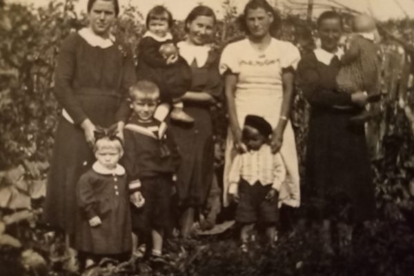 Czarno-białe zdjęcie rodzinne zrobione w ogrodzie. Na zdjęciu widocznych jest dziewięć osób – cztery dorosłe kobiety i pięcioro dzieci. Po lewej stronie stoi kobieta w ciemnej sukni z białym kołnierzykiem, przed nią dwójka dzieci – dziewczynka z dużą kokardą na głowie i chłopiec w marynarskim stroju. Obok stoi kobieta trzymająca dziecko na rękach, za nią kobieta w jasnej sukience, trzymająca chłopca ubranego w jasną kamizelkę i ciemne spodenki. Po prawej stronie kolejna kobieta trzymająca niemowlę. W tle gęsta roślinność, co sugeruje wiejski ogród. Zdjęcie ma charakter pamiątkowy, ukazując codzienne życie rodziny.
