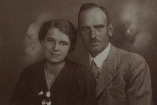 Czarno-białe portretowe zdjęcie przedstawia młode małżeństwo pozujące razem w studiu fotograficznym. Kobieta siedzi po lewej stronie, ma krótkie, starannie ułożone włosy i delikatny uśmiech. Ubrana jest w ciemną suknię z ozdobą z jasnych kwiatów przy dekolcie i nosi naszyjnik. Mężczyzna siedzi tuż obok niej, lekko za nią. Ma krótkie włosy zaczesane do tyłu, wąsy i spokojny wyraz twarzy. Ubrany jest w jasny, prążkowany garnitur z krawatem. Para patrzy prosto w obiektyw, emanując spokojem i bliskością. Tło jest rozmyte, typowe dla studyjnych portretów z lat 30. XX wieku.