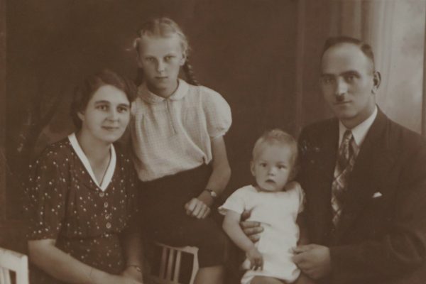 Czarno-białe zdjęcie portretowe z lat 40. XX wieku przedstawia czteroosobową rodzinę pozującą w studio fotograficznym. Po lewej stronie siedzi kobieta w ciemnej sukience w drobny wzór z jasnym kołnierzykiem. Ma krótkie, ciemne włosy i delikatny uśmiech. Obok niej stoi dziewczynka z jasnymi warkoczami, ubrana w jasną bluzkę i ciemną spódnicę – spogląda poważnie w stronę aparatu. Po prawej stronie zdjęcia siedzi mężczyzna w garniturze i krawacie, trzymający na kolanach małe dziecko ubrane w jasną sukienkę. Maluch ma jasne włosy i patrzy z zaciekawieniem w kierunku aparatu. Cała rodzina siedzi blisko siebie, emanując spokojem i bliskością. W tle widoczne są zasłony i rozmyte studio fotograficzne.