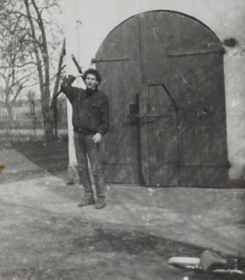 Czarno-białe zdjęcie przedstawia młodego mężczyznę stojącego na podwórzu przed dużą, drewnianą bramą z łukowym zwieńczeniem, która prowadzi do budynku gospodarczego. Mężczyzna ubrany jest w ciemną kurtkę i spodnie, na głowie ma czapkę. Uśmiecha się i trzyma w jednej ręce długi przedmiot – prawdopodobnie narzędzie rolnicze lub kij. Na pierwszym planie, przy krawędzi zdjęcia, leży przewrócony rower. W tle widać nagie drzewa, co sugeruje porę późnojesienną lub wczesnowiosenną.