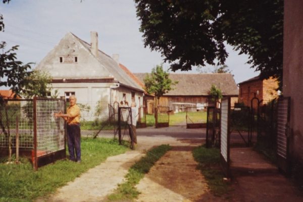 Kolorowe zdjęcie przedstawia wiejskie podwórze w słoneczny dzień. Na pierwszym planie po lewej stronie stoi starszy mężczyzna w pomarańczowej koszuli i ciemnych spodniach, trzymający metalową bramę wejściową. Po obu stronach widoczne są niskie ogrodzenia z siatki i przęsła z metalowymi ramami. W tle znajduje się jasny parterowy dom. W głębi widać jeszcze kilka budynków gospodarczych z czerwonej cegły i drewna. Podłoże to mieszanka trawy, ziemi i utwardzonych ścieżek. Zdjęcie pochodzi z lat 80. XX wieku.