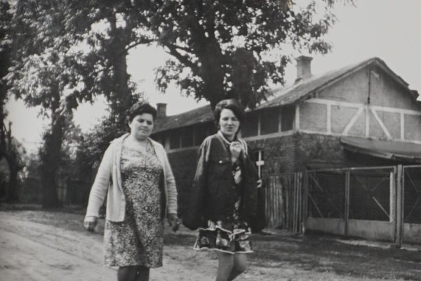 Czarno-białe zdjęcie przedstawia dwie kobiety idące wiejską, nieutwardzoną drogą. Kobieta po lewej stronie nosi sukienkę w kwiatowy wzór oraz rozpięty sweter, ma krótkie, ciemne włosy i stoi pewnie, spoglądając w stronę aparatu. Druga kobieta, po prawej, ubrana jest w krótki płaszcz i spódniczkę w kwiatowy wzór, z długimi ciemnymi włosami opadającymi na ramiona. W tle widoczny jest budynek o konstrukcji szachulcowej (drewniany szkielet wypełniony gliną lub cegłą), ogrodzenie z siatki i duże drzewa, co sugeruje wiejskie otoczenie. Zdjęcie oddaje codzienną scenę z życia wsi, prawdopodobnie z lat 60. lub 70. XX wieku.