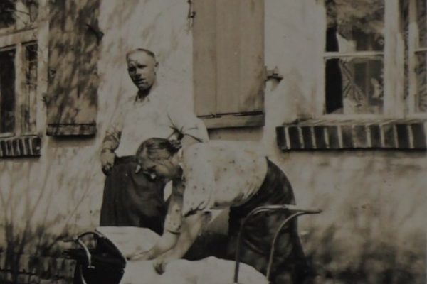 Czarno-białe zdjęcie przedstawia scenę z życia rodzinnego przed domem o tynkowanej ścianie i oknach z drewnianymi okiennicami. Na pierwszym planie stoi kobieta pochylona nad dziecięcym wózkiem, zaglądająca do jego wnętrza. Wózek jest duży, na czterech kołach, przykryty białym kocykiem. Obok kobiety stoi mężczyzna z ręką opartą na biodrze, patrzący w stronę aparatu. Ubrany jest w jasną koszulę i ciemne spodnie. Na twarzy ma spokojny wyraz. W tle widoczna jest fasada domu i fragmenty drzewa rzucające cień na ścianę. Zdjęcie emanuje ciepłem, codziennością i rodzinną atmosferą.