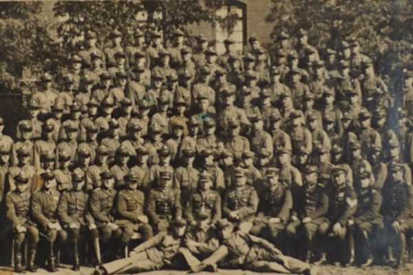 Czarno-białe zdjęcie przedstawia dużą grupę mężczyzn w wojskowych mundurach ustawionych do pamiątkowego zdjęcia grupowego. Żołnierze ustawieni są w wielu rzędach – niektórzy stoją, inni siedzą, a kilku leży z przodu na ziemi w pozycji zrelaksowanej. W pierwszym rzędzie siedzą oficerowie w bardziej ozdobnych mundurach z odznaczeniami, a za nimi szeregowi żołnierze. W tle widoczne są drzewa i ceglana fasada budynku z dużymi oknami, co sugeruje, że zdjęcie zostało wykonane na dziedzińcu koszar lub szkoły wojskowej. Zdjęcie emanuje dyscypliną i dumą z przynależności do formacji wojskowej.