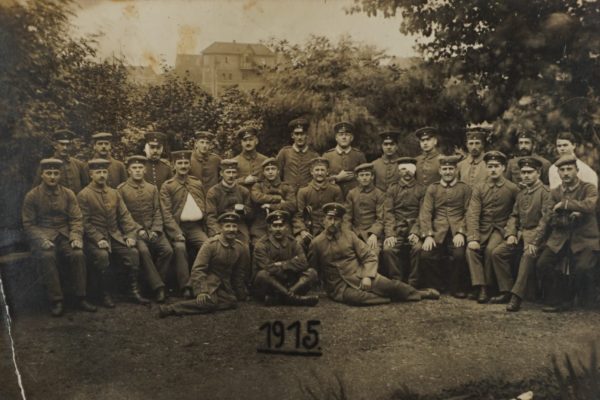 Czarno-białe zdjęcie grupowe z 1915 roku przedstawia około trzydziestu mężczyzn w wojskowych mundurach z czasów I wojny światowej. Ustawieni są w trzech rzędach – część siedzi, część stoi, a kilku siedzi na ziemi z przodu. Większość z nich nosi czapki z daszkiem, niektórzy mają wąsy. W tle widoczne są drzewa i zabudowania, co sugeruje, że zdjęcie zostało wykonane na terenie parku lub ogrodu, prawdopodobnie w mieście. Atmosfera zdjęcia jest poważna, lecz nie ponura – to portret pamiątkowy grupy żołnierzy.