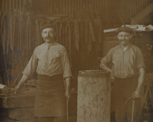 Stare, sepiowe zdjęcie przedstawia dwóch mężczyzn w kuźni. Obaj mają wąsy, noszą robocze, zakasane koszule, spodnie i nakrycia głowy — charakterystyczne dla kowali z przełomu XIX i XX wieku. Mężczyzna po lewej stronie trzyma metalowy pręt i stoi przy kowadle, u jego pasa wisi fartuch ochronny. Po prawej stronie drugi kowal opiera rękę na dużym drewnianym pniu i trzyma ciężki młot lub szczypce kowalskie. W tle widoczne są różnego rodzaju narzędzia zawieszone na ścianie: młoty, obcęgi, pilniki, a także inne kowalskie wyposażenie. Całość wnętrza kuźni sprawia wrażenie surowości i tradycyjnego rzemiosła.