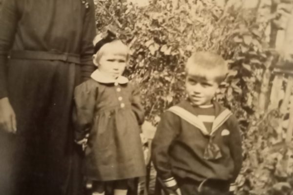 Czarno-białe zdjęcie przedstawia kobietę i dwoje dzieci w ogrodzie. Kobieta stoi po lewej stronie, ubrana w ciemną suknię z białym kołnierzykiem i przypinkami na piersi, z ciemnymi włosami zaczesanymi do tyłu. Obok niej stoi dziewczynka w ciemnej sukience z białym kołnierzykiem i kokardą we włosach, ustawiona na małym krzesełku. Obok niej stoi chłopiec ubrany w strój marynarski z krótkimi spodenkami, marynarskim kołnierzem i krawatem. W tle widoczna jest bujna roślinność oraz ogrodzenie, co sugeruje, że zdjęcie wykonano w ogródku przydomowym. Atmosfera zdjęcia jest spokojna i domowa.