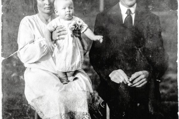Czarno-białe, lekko uszkodzone zdjęcie z około 1930 roku przedstawia trzyosobową rodzinę siedzącą na krzesłach na świeżym powietrzu. Po lewej stronie siedzi kobieta w jasnej sukni z koronkowym wykończeniem – trzyma na kolanach małe dziecko ubrane w jasną sukienkę i buciki. Dziecko ma poważny wyraz twarzy i spogląda lekko w bok. Po prawej stronie siedzi mężczyzna w ciemnym garniturze z białą koszulą i krawatem, z uśmiechem na twarzy i splecionymi dłońmi. W tle rozmyte drzewa i trawa, co sugeruje, że zdjęcie wykonano w ogrodzie lub na podwórzu. Fotografia ma ślady zagięć i przetarć, co podkreśla jej wiek i charakter pamiątkowy.