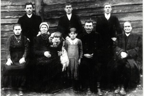 Czarno-białe, archiwalne zdjęcie grupowe przedstawiające wielopokoleniową rodzinę, ustawioną przed drewnianą ścianą budynku (najprawdopodobniej chaty wiejskiej). Łącznie na zdjęciu jest 11 osób – 6 mężczyzn i 5 kobiet, w tym dwie dziewczynki. Wszyscy są ubrani w odświętne stroje z epoki – kobiety mają ciemne suknie i korale, a mężczyźni garnitury lub długie płaszcze. Z przodu siedzą starsze osoby i dzieci, z tyłu stoją młodsi mężczyźni. Po prawej stronie zdjęcia siedzi starszy mężczyzna z siwymi włosami – wygląda na nestora rodu. Scena jest statyczna, a wyraz twarzy wszystkich osób jest poważny i skupiony – typowe dla fotografii z początku XX wieku. Zdjęcie ma wyraźny klimat dokumentalny i rodzinny.