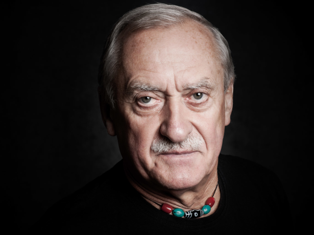 Na zdjęciu znajduje się postać starszego mężczyzny. Ma on siwe włosy oraz wąsy. Ubrany jest w sweter koloru czarnego. Na szyi ma założony rzemyk z czerwonymi, turkusowymi i czarnymi koralikami.. Za nim tło w jednolitym czarnym kolorze.