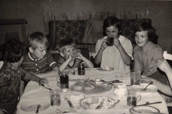 Zdjęcie czarno-białe przedstawiające grupę pięciorga dzieci siedzących przy stole nakrytym obrusem. Na stole stoją szklanki w metalowych koszyczkach, talerze, pieczywo i kawałki ciasta. Dzieci rozmawiają, piją i śmieją się, tworząc ciepłą, rodzinną atmosferę. W tle widać kanapę przykrytą narzutą.