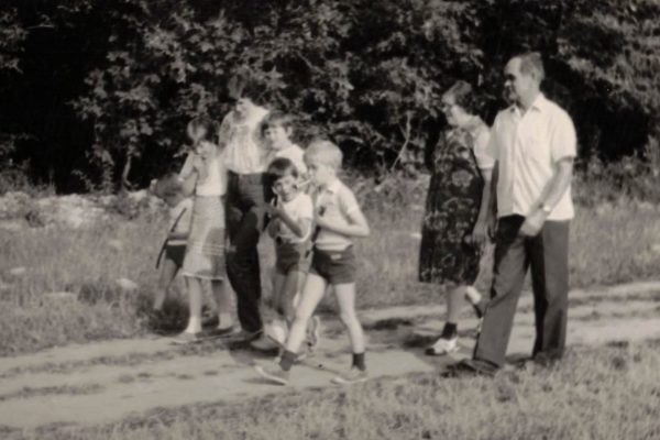 Czarno-białe zdjęcie przedstawia grupę osób spacerujących polną drogą w pobliżu lasu. Na przedzie idą dwaj chłopcy w krótkich spodenkach, za nimi kolejne dzieci oraz dwoje dorosłych – kobieta w sukience i mężczyzna w jasnej koszuli. Cała grupa porusza się razem, tworząc pogodną, rodzinną scenę letniego spaceru.