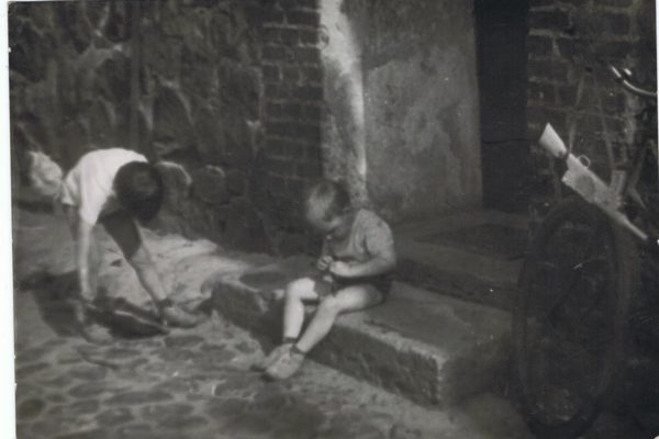 Czarno-białe zdjęcie przedstawia dwóch chłopców bawiących się przed wejściem do budynku z cegły i kamienia. Jeden chłopiec stoi tyłem, z zabawką przewieszoną przez ramię, drugi siedzi na progu i patrzy w dół. Obok, przy ścianie, oparty jest rower. Atmosfera uchwycona na fotografii oddaje codzienność dzieciństwa lat 70.