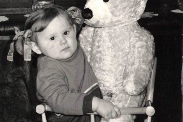 Czarno-białe zdjęcie przedstawiające małe dziecko siedzące na krześle obok dużego pluszowego misia. Dziewczynka ubrana w sweter i spodnie, z kokardkami we włosach, patrzy w stronę aparatu.