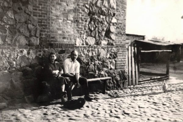 „Czarno-białe zdjęcie przedstawiające dwie osoby siedzące na ławce przy kamienno-ceglanej ścianie budynku. Obok widać brukowaną uliczkę i drewniane ogrodzenie, a w oddali stoi pojedynczy ptak.”