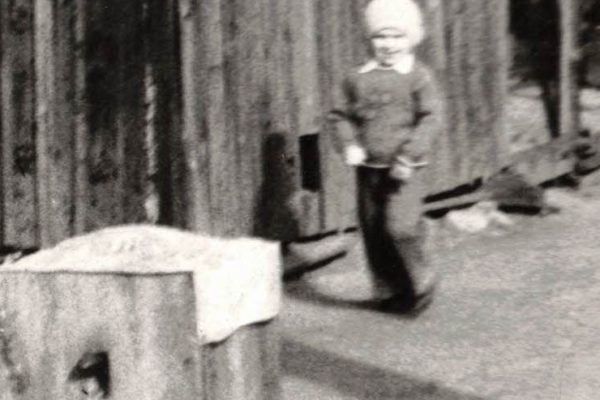 Czarno-białe zdjęcie przedstawiające małe dziecko w czapce stojące obok drewnianego budynku o szorstkich deskach, prawdopodobnie kurnika. Na pierwszym planie widać duży drewniany pień lub skrzynię przykrytą materiałem