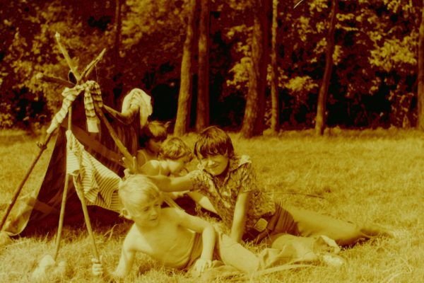 Fotografia w odcieniach sepii przedstawiająca grupę dzieci bawiących się na łące obok prowizorycznego namiotu z patyków i koców. Na pierwszym planie chłopiec bez koszulki leży w trawie, a obok niego drugi chłopiec w koszuli w paski uśmiecha się i pokazuje mu dwa palce nad głową. W tle widać kolejne dzieci oraz drzewa.