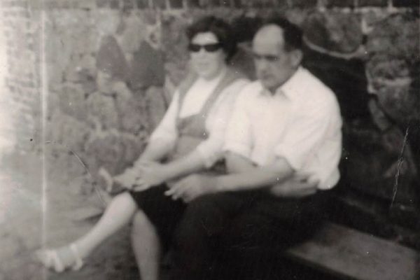 Czarno-białe, lekko rozmyte zdjęcie przedstawiające mężczyznę i kobietę siedzących razem na ławce przy kamiennym murze. Kobieta w sukience i okularach przeciwsłonecznych, mężczyzna w białej koszuli z podwiniętymi rękawami. Trzymają się blisko siebie, tworząc spokojną, intymną scenę