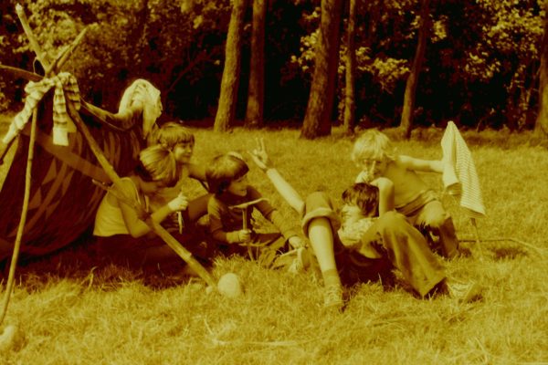 „Zdjęcie w odcieniach sepii przedstawiające grupę dzieci bawiących się na łące obok prowizorycznego namiotu z patyków i koca. Część dzieci siedzi na trawie i śmieje się, jedno dziecko trzyma kij, a inne pokazuje ręką znak w górze. W tle widoczne drzewa.”