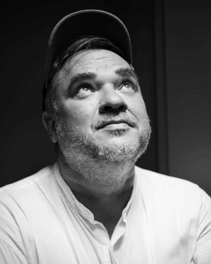 Fotografia przedstawia autora książek dla dzieci Grzegorza Kasdepkę. Mężczyzna w ujęciu od klatki piersiowej w górę. Ubrany jest w biała koszule, a na głowie ma czapkę z daszkiem. Spogląda w górę. Zdjęcie jest czarno-białe.