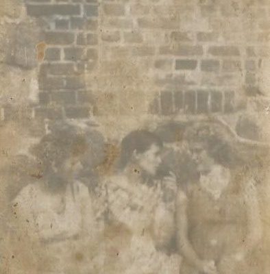 Stare, wyblakłe czarno-białe zdjęcie przedstawiające trzy młode kobiety stojące przed ceglanym murem. Fotografia jest mocno zniszczona i rozmazana, ale widać, że kobiety stoją blisko siebie i rozmawiają.