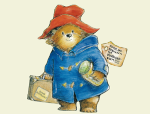 Ilustracja przedstawia niedźwiedzia ubranego w niebieski płaszcz. Na głowie ma czerwony kapelusz. W prawej swej łapie trzyma walizkę, a w lewej słoik z marmoladą. Na szyi ma zawieszoną informację z napisem proszę zaopiekować się tym niedźwiadkiem dziękuję.