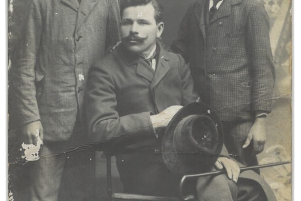 Trzech mężczyzn na zabytkowej fotografii studyjnej z początku XX wieku – siedzący dorosły mężczyzna z kapeluszem oraz dwóch chłopców w eleganckich garniturach.