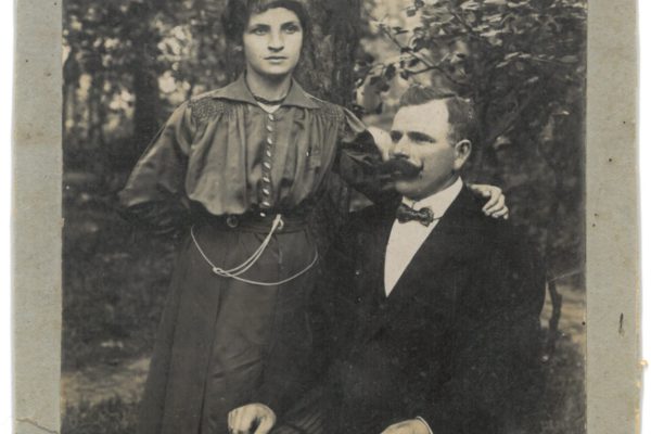 Czarno-biała, archiwalna fotografia przedstawiająca kobietę stojącą obok siedzącego mężczyzny w plenerze, prawdopodobnie w lesie lub parku. Kobieta ubrana jest w długą suknię z długimi rękawami, ma krótkie, falowane włosy i opiera dłoń na ramieniu mężczyzny. Mężczyzna siedzi na krześle, ubrany w elegancki garnitur z muszką, ma charakterystyczne wąsy. Oboje patrzą poważnie w stronę aparatu. Zdjęcie ma jasną kartonową ramkę i nosi ślady wieku.