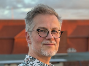 Zdjęcie przedstawia twarz mężczyzny. Ma on siwe, krótkie włosy oraz bródkę z niewielkim wąsem. Na nosie okulary w czarnych oprawkach. W tle widać nieco rozmazane, pomarańczowe budynki.